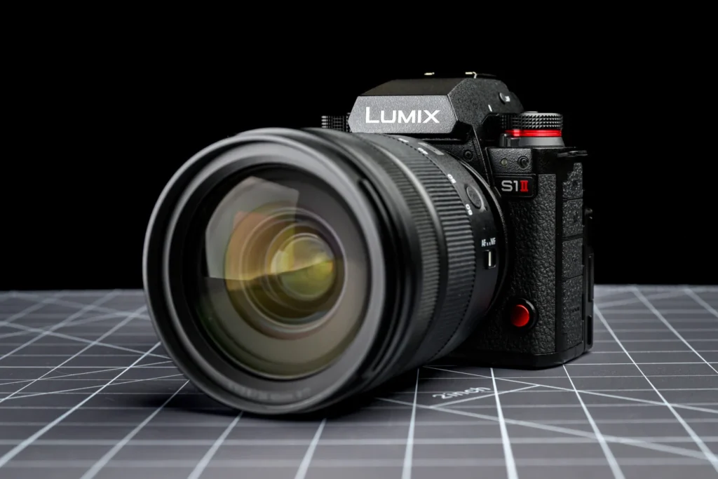 Panasonic Lumix S1 II 카메라와 대형 렌즈가 격자무늬 배경 위에 놓여 있는 모습. 카메라 전면 디자인과 LUMIX 로고가 강조되어 있다.