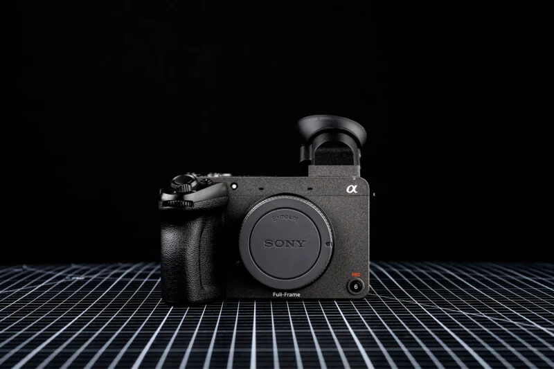 검은색 배경과 격자무늬 바닥 위에 놓인 Sony FX2 풀프레임 시네마 카메라. 렌즈가 장착되지 않은 상태로 바디캡이 장착되어 있으며, 전자식 뷰파인더가 상단에 돌출되어 있다.
