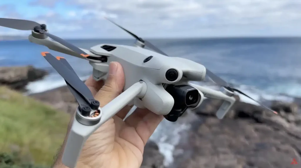 DJI Mini 4 Pro 드론을 손에 들고 있는 모습. 바닷가 절벽 위에서 촬영된 장면으로, 드론의 렌즈와 회전 중인 프로펠러가 강조되어 있다.