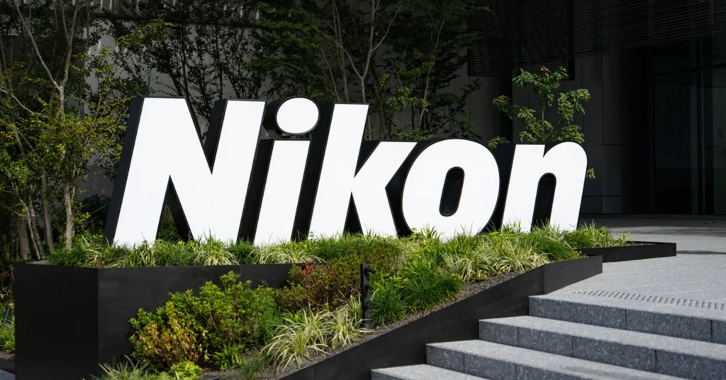 니콘(Nikon) 본사 앞에 설치된 대형 로고 조형물. 푸른 식물과 나무들이 둘러싸인 현대적인 건물 외관 앞에 하얀색 'Nikon' 로고가 선명하게 보인다.