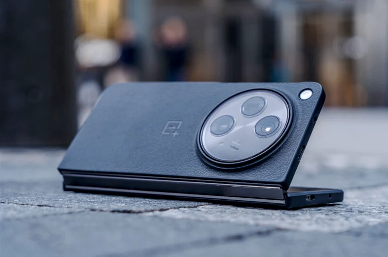 반쯤 펼쳐진 상태의 OnePlus Open 스마트폰이 콘크리트 바닥 위에 놓여 있고, 원형 카메라 모듈이 강조되어 있다.