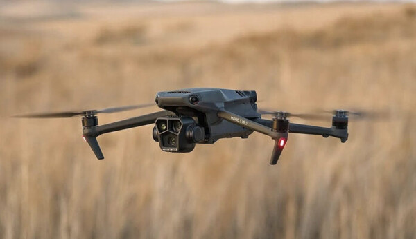 넓은 황금빛 들판 위를 비행 중인 DJI Mavic 4 Pro 드론. 전면에 고성능 메인 카메라와 망원 카메라 모듈이 장착되어 있고, 프로펠러가 회전하고 있는 모습.