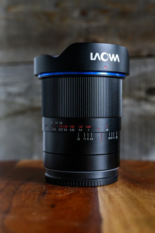 Laowa 15mm f/4.5 Wide Angle Macro 렌즈가 나무 테이블 위에 세워져 있으며, 금속 재질의 렌즈 바디와 빨간 초점 거리, 흰색 조리개 수치가 각인된 모습이 보인다. 블루 링과 LAOWA 로고가 인쇄된 렌즈 후드가 장착되어 있다.