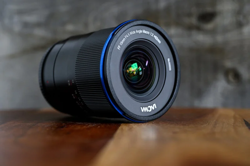 Laowa 15mm f/4.5 렌즈가 테이블 위에 비스듬히 놓여 있으며, 전면에는 “FF 15mm f/4.5 Wide Angle Macro 1:2 ø62mm” 문구가 선명하게 각인되어 있다. 렌즈 내부 유리에 빛 반사가 보이며, 고급스러운 광학 유리 느낌을 전달한다.
