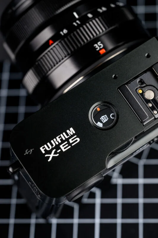 Fujifilm X-E5 상단부 로고와 셔터 모드 다이얼 클로즈업, 35mm 렌즈 장착된 상태