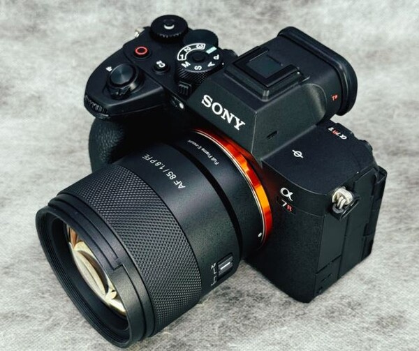 소니 A7R V 미러리스 카메라에 삼양 Prima AF 85mm F1.8 FE 렌즈가 장착되어 있는 모습. 카메라와 렌즈 모두 검은색이며, 렌즈에는 "AF 85 / 1.8 FE"와 "Full Frame E-mount" 문구가 새겨져 있다. 카메라의 렌즈 마운트 부분에는 주황색 링이 보인다. 전체적으로 회색의 질감 있는 배경 위에 놓여 있다.