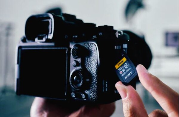 Sony "TOUGH" CFexpress Type A 메모리 카드를 들고 있으며, 흐릿하게 처리된 Sony Alpha 시리즈 카메라의 메모리 카드 슬롯 근처에 위치해 있습니다. 카드의 용량과 속도 정보, Sony 로고가 보입니다.