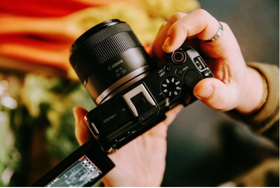 두 손이 Canon EOS R50V 카메라를 잡고 있습니다. 카메라는 렌즈가 위쪽을 향하도록 약간 기울어져 있으며, LCD 화면이 옆으로 펼쳐져 있습니다. 카메라의 버튼과 다이얼, 렌즈의 모델명이 보입니다. 배경은 흐릿하게 처리되어 카메라에 초점이 맞춰져 있습니다.