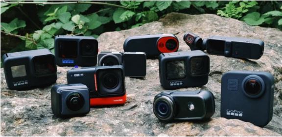 다양한 액션 카메라와 짐벌 카메라들이 울퉁불퉁한 바위 위에 놓여 있습니다. 전면에 GoPro Hero 시리즈, Insta360 One R, DJI Osmo Pocket 등 여러 브랜드의 카메라들이 모여 있어 액션 카메라 시장의 다양한 제품군을 보여줍니다. 배경에는 녹색 식물이 보입니다.
