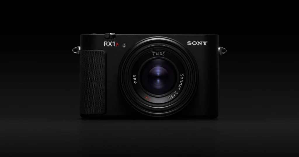 전면에서 바라본 소니 RX1R III 카메라의 모습입니다. 렌즈는 중앙에 위치하며, 그 위로 "SONY" 로고가 새겨져 있습니다. 카메라 왼쪽 상단에는 "RX1R III" 로고가 빨간색 'R' 강조와 함께 보입니다. 렌즈 주변에는 초점 거리와 조리개 값이 표시되어 있습니다.