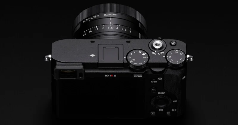 소니 RX1R III 카메라의 윗면을 보여주는 사진입니다. 렌즈의 초점 거리 조절링과 조리개 조절링이 뚜렷하게 보입니다. 카메라 상단에는 다양한 모드 다이얼, 셔터 버튼, 전원 스위치, 사용자 지정 버튼(C1) 등이 배치되어 있습니다. 팝업 뷰파인더가 내려가 있는 모습도 확인할 수 있습니다.