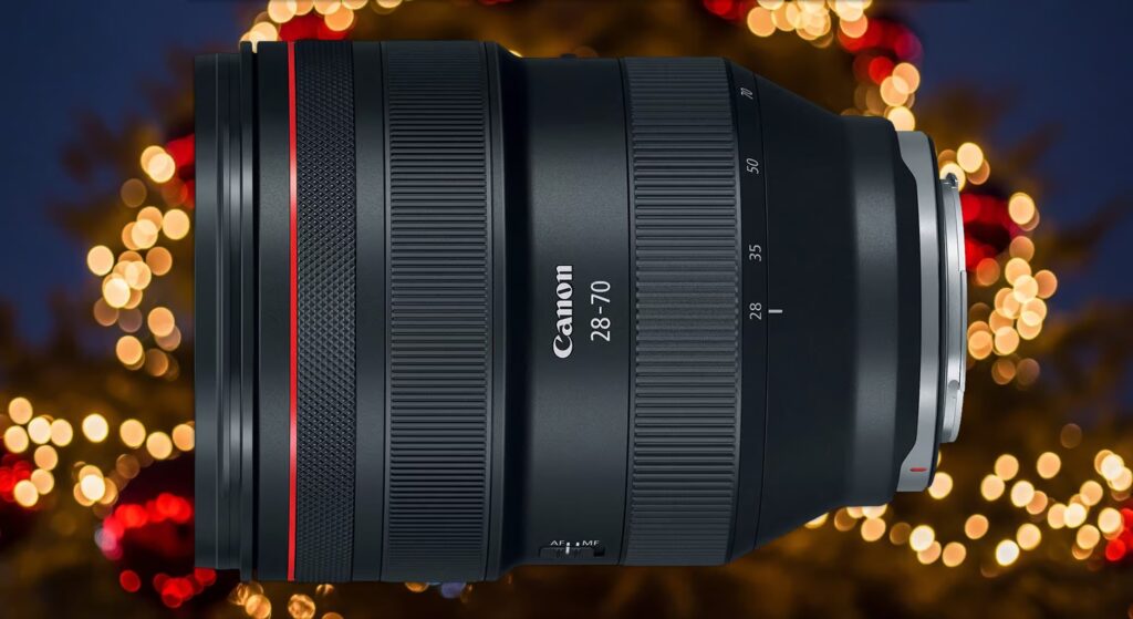 Canon RF 28-70mm 렌즈가 크리스마스 트리처럼 보이는 흐릿한 배경의 노란색과 빨간색 보케 조명 위에 놓여 있습니다. 렌즈는 검은색이며, 중앙에 "Canon 28-70"이라고 쓰여 있고, 빨간색 링과 초점 거리 표시(28, 35, 50, 70)가 보입니다. 렌즈 아래쪽에는 AF/MF 스위치가 있습니다.