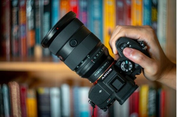 "책장 앞에서 Sony Alpha 미러리스 카메라를 한 손으로 들고 있는 모습. 장착된 렌즈는 고급 줌렌즈로 보인다."