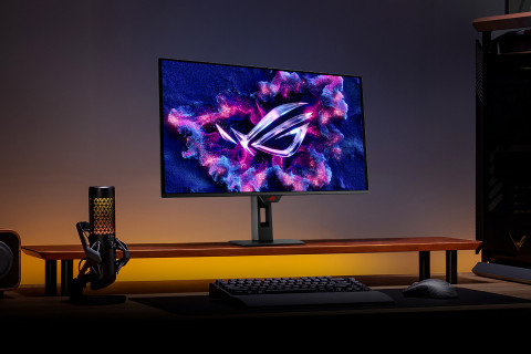 어두운 환경의 게이밍 데스크에 놓인 ASUS ROG 게이밍 모니터. 모니터 화면에 ROG 로고가 선명하게 빛나고 있으며, 옆에는 RGB 마이크와 키보드, 마우스가 배치되어 있어 몰입감 있는 게이밍 분위기를 연출한다.