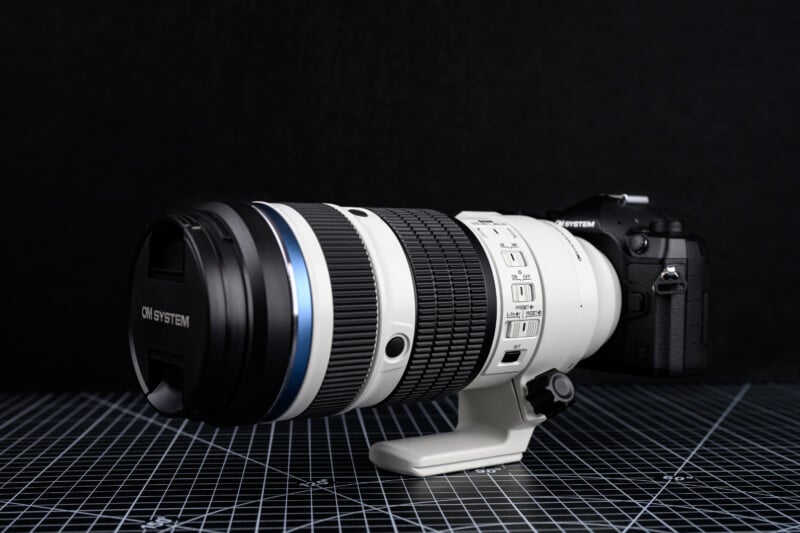 OM System M.Zuiko 50-200mm f/2.8 IS Pro 렌즈가 카메라 바디에 장착된 상태로 삼각대 마운트 위에 놓여 있는 모습, 흰색 바디와 블루 링이 돋보임