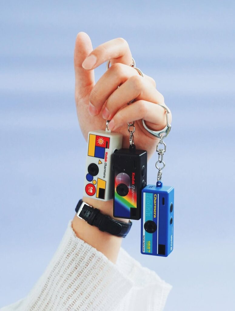 손에 들린 세 개의 코닥 차메라(Kodak Charmera) 키링 디지털카메라. 왼쪽은 흰색 바디에 원색 기하학 무늬 디자인, 가운데는 검은색 바디에 무지개 패턴, 오른쪽은 파란색 바디에 ‘1997’ 레트로 라벨이 붙어 있다.