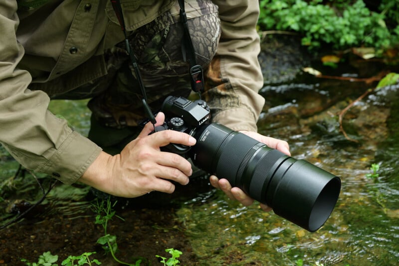야외 숲 속 개울가에서 카무플라주 재킷을 입은 사진가가 LUMIX 카메라와 망원 줌렌즈를 손에 들고 촬영 준비하는 장면.
