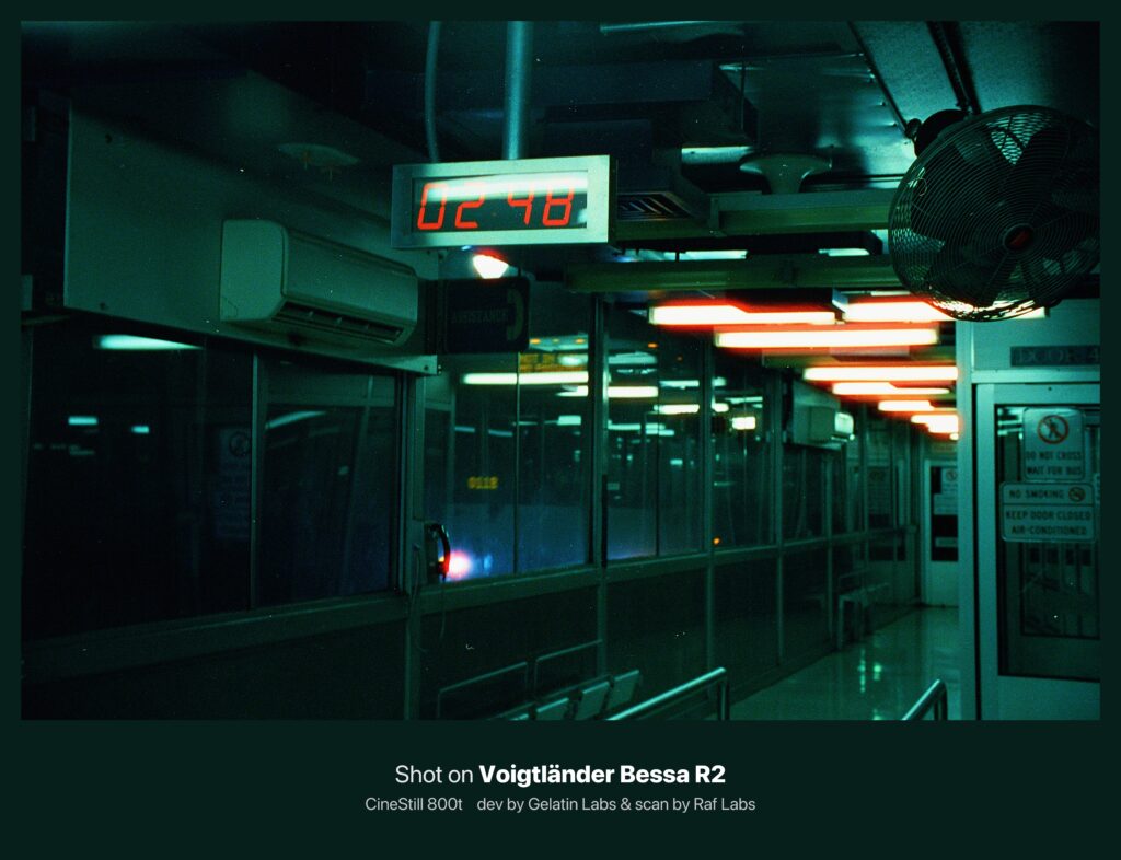 어두운 실내 통로의 풍경. 천장에 붉은 형광등이 줄지어 켜져 있으며, 전광판에는 숫자가 표시되고 있다. 푸른빛과 붉은빛이 뒤섞여 사이버펑크적 분위기를 자아내며, 필름 특유의 강한 색 번짐과 입자가 강조된다.