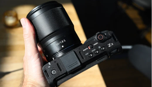 손에 들린 니콘 ZR 시네마 카메라 본체. 50mm f/1.8 S 렌즈가 장착되어 있으며, 상단에는 ‘ZR’ 로고와 촬영 모드 전환 버튼, 녹화 버튼 등이 보인다.