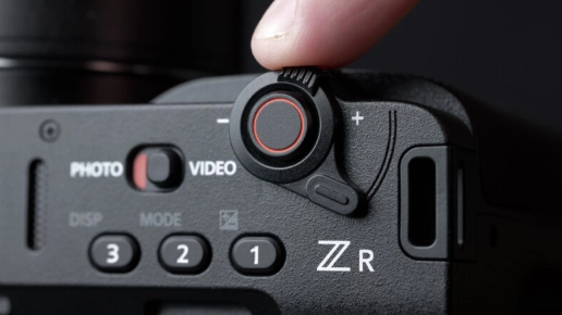 손가락이 니콘 ZR의 상단 줌 레버를 조작하는 클로즈업. 옆에는 PHOTO/VIDEO 전환 스위치와 붉은 원형 버튼, ZR 로고가 보인다.