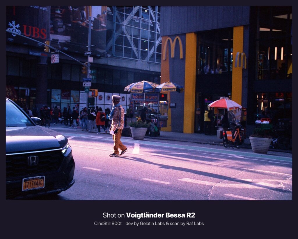 맨해튼 거리에서 횡단보도를 건너는 인물. 배경에는 맥도날드 매장과 노란 간판, 핫도그 노점이 보이며, 군중이 모여 있다. 강한 햇살과 네온빛이 섞이며 필름 특유의 붉고 보랏빛 할레이션이 번진다.