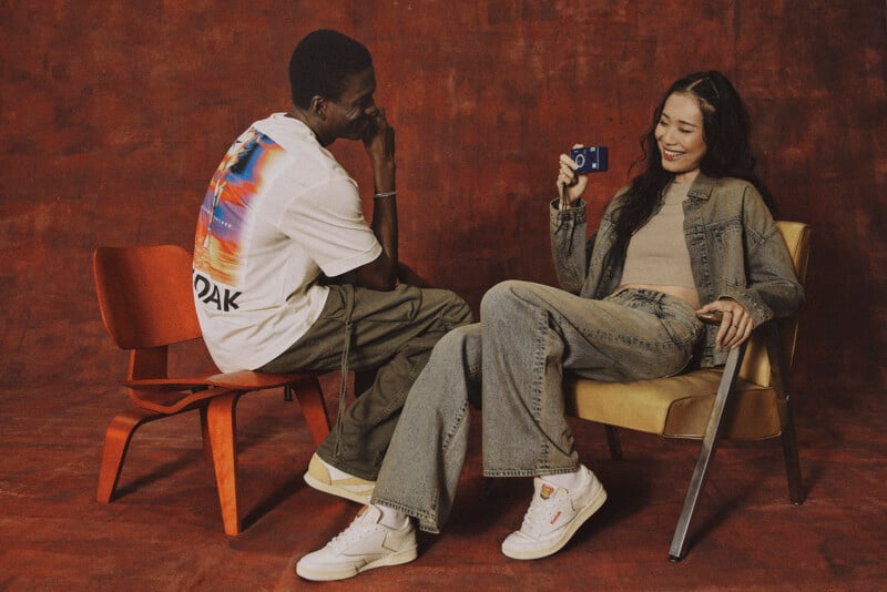 코닥 그래픽 티셔츠와 캐주얼 팬츠를 착용한 남성 모델과 데님 재킷과 청바지를 착용한 여성 모델이 붉은 배경의 스튜디오에서 Kodak 카메라를 들고 웃고 있는 모습