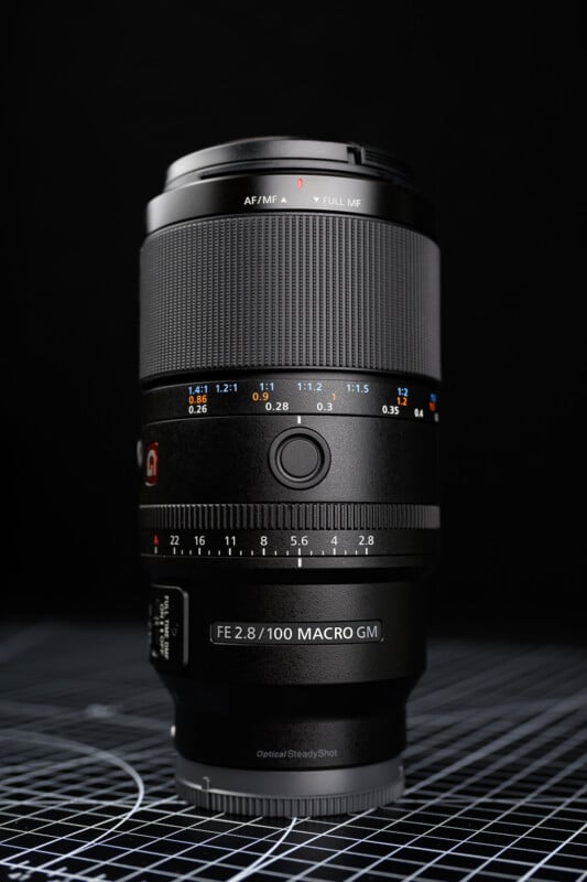 검은 배경 위에 세워진 소니 FE 100mm f/2.8 매크로 G 마스터 렌즈.
조리개 수치와 초점 거리 표시가 선명하게 보이며, 견고한 메탈 바디의 질감이 강조되어 있다.