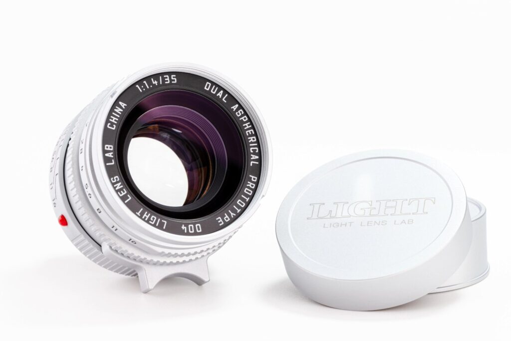 Light Lens Lab 35mm f/1.4 Aspherical “11873” 렌즈의 정면 이미지. 전면부에는 ‘DUAL ASPHERICAL PROTOTYPE’ 각인이 있으며, 옆에는 Light Lens Lab 로고가 새겨진 금속 캡이 함께 놓여 있다. 렌즈의 고급스러운 광택과 정교한 구조가 돋보인다.
