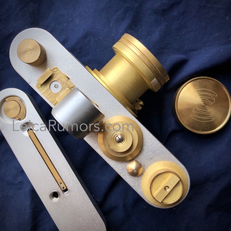 카메라 하단 커버가 분리된 상태로 놓여 있는 바르낙 레플리카 필름 카메라. 황동으로 제작된 렌즈 마운트와 조작 다이얼이 강조되어 있으며, 옆에는 렌즈 캡도 함께 배치되어 있다.