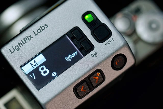 LightPix Labs FlashQ FM2의 상단 조작부를 근접 촬영한 이미지. OLED 디스플레이에 M 모드와 출력 수치가 표시되어 있으며, TTL·무선 아이콘과 버튼 조작부가 보인다. 금속 재질의 표면과 깔끔한 인터페이스가 돋보인다.
