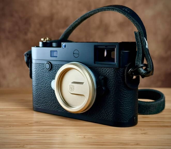갈색 배경과 나무 테이블 위에 블랙 가죽 스트랩이 연결된 라이카 M 카메라가 놓여 있다. 전면에는 금색 Owlkrown 웨더실드 메탈 바디캡이 장착되어 있으며, 고급스러운 질감과 클래식한 디자인이 강조되어 있다.
