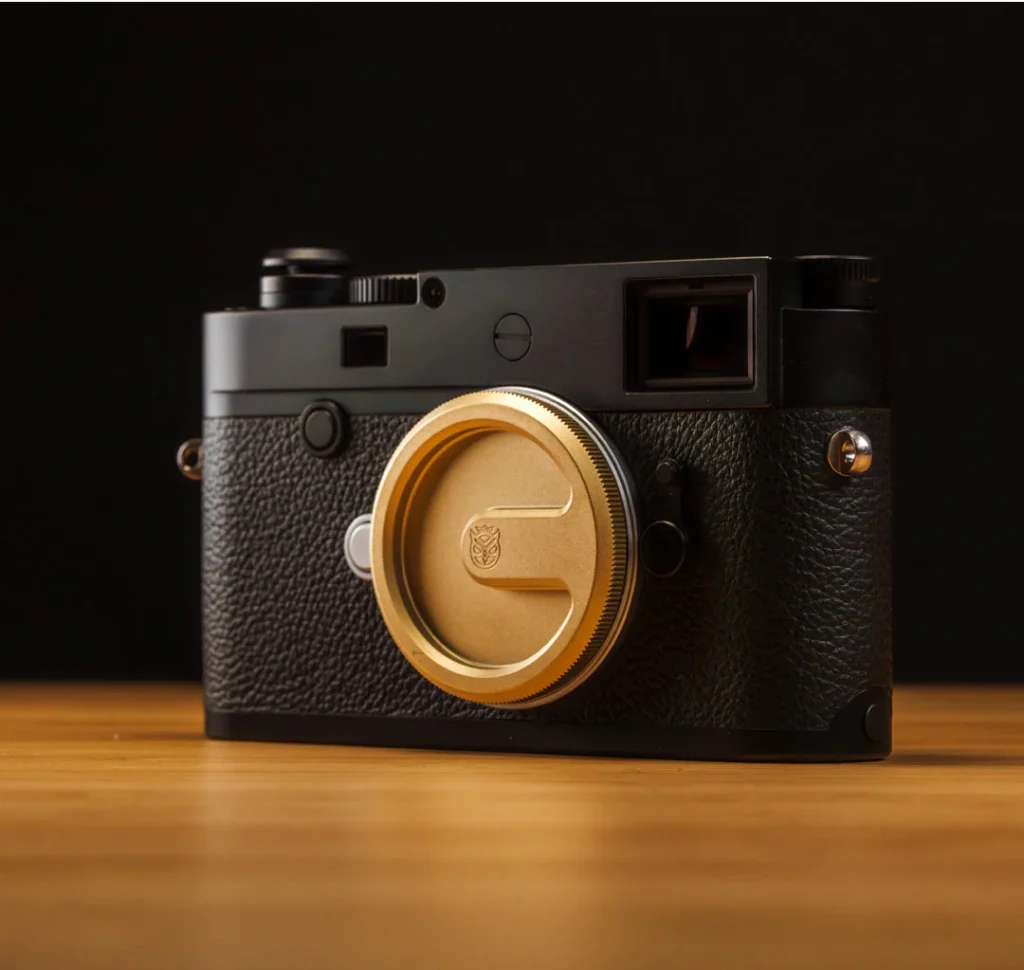 검은색 배경과 나무 테이블 위에 놓인 라이카 M 카메라. 전면에는 황동 소재의 Owlkrown 웨더실드 메탈 바디캡이 장착되어 있으며, 매트한 질감과 정밀 가공된 표면이 조명을 받아 은은하게 반사되고 있다.