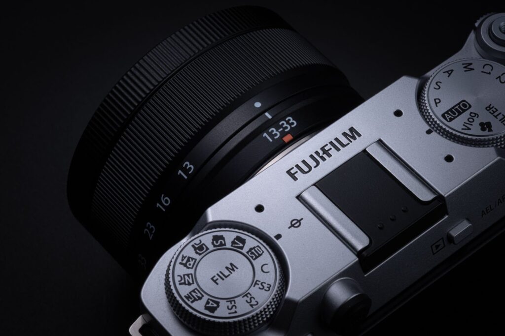 후지필름 X 시리즈 실버 바디 카메라(X-T30 III 예상)에 장착된 XC 13-33mm f/3.5-6.3 OIS 렌즈의 상단 근접 이미지.
카메라의 다이얼과 ‘FILM’ 모드 표시, 렌즈의 13-33mm 줌 표시가 뚜렷하게 보인다.