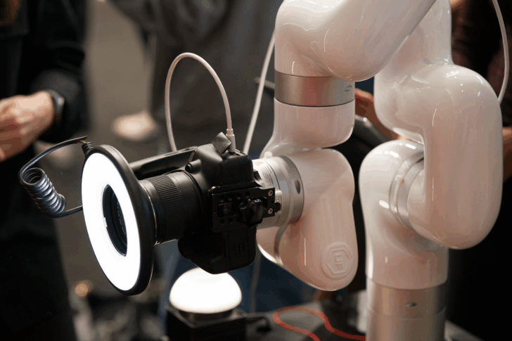 하얀색 로봇 팔에 장착된 캐논 카메라가 링 조명을 켜고 인물 촬영을 준비하는 모습. Salon de la Photo 2025 행사장에서 관람객들이 주변에서 지켜보고 있다.