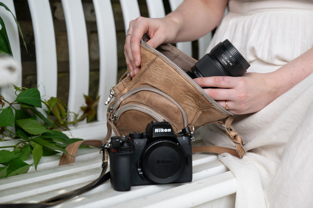 흰색 원피스를 입은 여성이 벤치에 앉아 가죽 가방에서 니콘 NIKKOR Z DX MC 35mm f/1.7 렌즈를 꺼내고 있다. 옆에는 니콘 Z 시리즈 카메라가 놓여 있으며, 주변에는 녹색 식물 잎이 보인다.