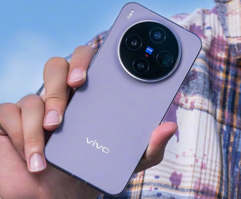 보라색 Vivo X300 Pro 스마트폰을 손에 든 사람의 모습. 원형 Zeiss 트리플 카메라 모듈이 후면 중앙 상단에 배치되어 있으며, 배경은 푸른 하늘과 체크무늬 셔츠가 어우러져 있다.